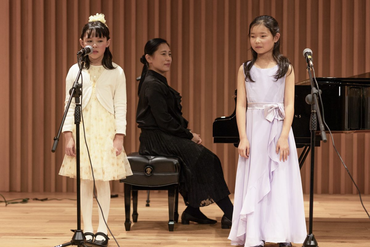 Children singing at the microphones with their teacher accompanying on piano