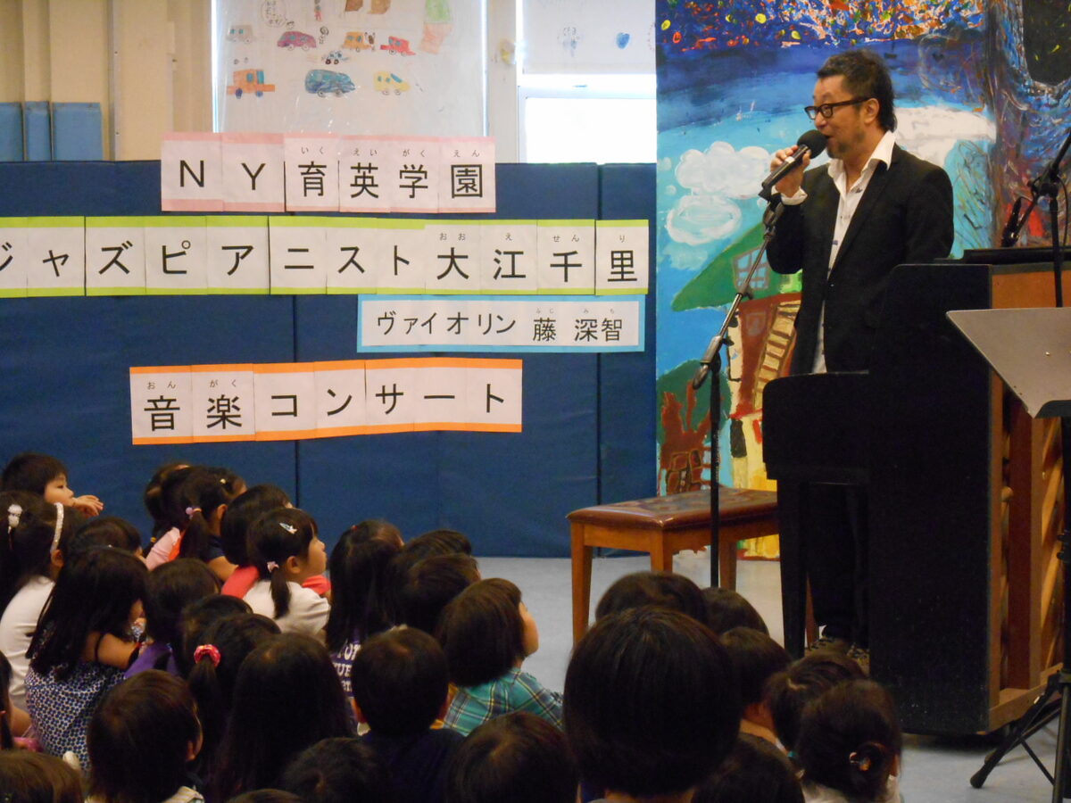 「NY育英学園 ジャズピアニスト大江千里 ヴァイオリン 藤深智 音楽コンサート」の掲示の前で、マイクを手に話す大江千里さん